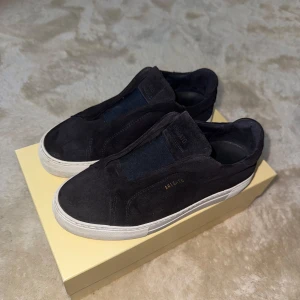 Mörkblå Axel Arigato Laceless 360 - Stilrena svarta slip-on sneakers från Axel Arigato i mocka med vit platt sula. Skorna har minimalistisk design utan snörning och diskret guldfärgad logga på sidan. Perfekta för dig som gillar en clean och modern look. Säljer pga utväxt. Skick 9/10, nypris: 2700 kr. Påse samt box medföljer.