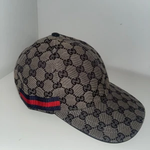 Gucci keps med GG-mönster och röd-blå rand - Snygg keps från Gucci i beige med klassiskt GG-monogram över hela kepsen. Har en röd och blå rand på sidan och justerbar rem i mörkblått skinn baktill. Perfekt för dig som vill sticka ut med en lyxig accessoar.