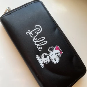 Svart plånbok med Belle-tryck - Plånbok i svart syntetmaterial med söt Belle-illustration och text på framsidan. Har dragkedja runtom, flera kortfack och ett myntfack med dragkedja inuti. Perfekt för dig som gillar gulliga detaljer och vill ha ordning på dina grejer.