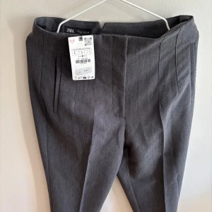Grå kostymbyxor från Zara, stl L - Snygga grå kostymbyxor från Zara i storlek L. Byxorna har en klassisk rak passform med pressveck och diskreta fickor framtill. Perfekta för en clean och stilren look. Materialet känns mjukt och följsamt.