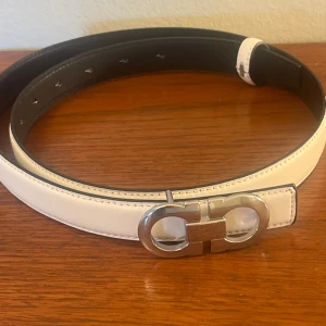 Vitt bälte Ferragamo - Snyggt vitt bälte från Salvatore Ferragamo i skinn med en ikonisk silverfärgad dubbel-Gancini-spänne. Bältet har stilrena sömmar och en klassisk, rak form. Perfekt för att lyfta din outfit med en exklusiv touch. Min leverantör säger att den är äkta, men jag tvekar därför det låga priset. det är kvinno modellen det innebär att det är lite mindre/smalare.