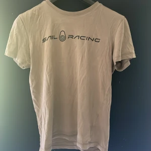 Beige t-shirt från Sail Racing S - Snygg beige t-shirt från Sail Racing med svart logga tryckt på bröstet. Klassisk rund hals och korta ärmar. Skön passform och tillverkad i mjuk bomull, perfekt till jeans eller shorts. Nypris 400:-