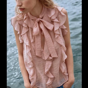 Rosa/peach volangblus med knyt från Zara - Supersöt ljusrosa blus från Zara med volanger längs axlar och framstycke. Blusen är ärmlös och har en snygg knytning vid halsen. Perfekt att styla med jeans eller kjol! Storleken är XS men hade sagt att det passar både XS och S. Prislappen e kvar då tröjan aldrig kom till användning! Hör av er vid frågor!