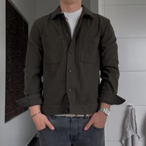 Olivgrön overshirt från These Glory Days - Snygg olivgrön overshirt från These Glory Days med två stora bröstfickor och knäppning framtill. Jackan har en rak passform och är tillverkad i ett mjukt bomullstyg, perfekt att slänga över en t-shirt. Stilren och enkel design som funkar året runt.