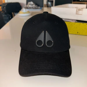 Svart keps från Moose Knuckles - Snygg svart keps från Moose Knuckles med deras ikoniska metallogo framtill. Kepsen har böjd skärm, justerbart spänne bak och är tillverkad i bomull. Perfekt accessoar för en clean och stilren look.