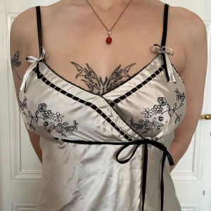 Omlottklänning med broderi - Snygg omlottklänning i satin med svarta axelband och knyt i midjan. Klänningen har broderade svarta blommor, små rosetter vid axelbanden och en v-ringad omlottringning. Perfekt för dig som gillar romantiska detaljer och en lyxig känsla.