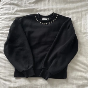 Svart sweatshirt med pärlor Kappahl XS - Svart sweatshirt från Kappahl i storlek XS med rund halsringning och snygga pärldetaljer längs kragen. Tröjan har långa ärmar och ribbade muddar vid ärmslut och nederkant. Perfekt för dig som vill ha en basic tröja med lite extra stil.