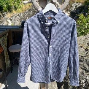 Blårutig skjorta från Morris - Skjorta från Morris med button down-krage och vita knappar. Skjortan har lång ärm och broderad logga på bröstet. Tillverkad i mjuk bomull. Inga defekter.