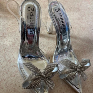 Glittriga pumps med rosett från Clubaldo , köpt den i utomland , har använt den en gång. - Snygga transparenta pumps från Clubaldo med spetsig tå och låg klack. Skorna har en glittrig rosett framtill och är i silverfärgad syntet med plastdetaljer. Perfekta för dig som vill sticka ut med en elegant och trendig look.