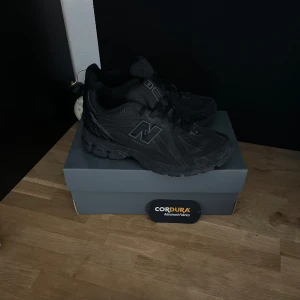 Svarta New Balance sneakers Cordura - Snygga helsvarta New Balance sneakers med robust design och detaljer i Cordura-tyg för extra slitstyrka. Skorna har en chunky sula, reflekterande detaljer och klassisk N-logga på sidan. Perfekta för dig som gillar streetwear och vill ha något tåligt och stilrent.