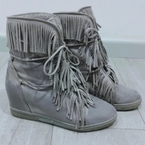 Grå mockaboots med fransar - Snygga grå boots i mocka med coola fransdetaljer och snörning framtill. Skorna har en diskret kilklack som ger extra höjd utan att tumma på komforten. Perfekta för dig som vill sticka ut med en boho-inspirerad stil.