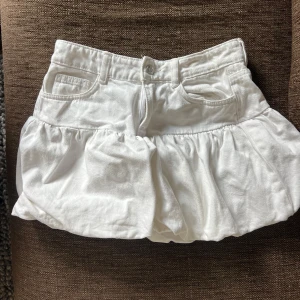 Vit bubblig kortkjol med fickor - Supersöt vit kortkjol med bubblig form och bred linning. Kjolen har fickor framtill och bälteshällor, vilket ger en cool och avslappnad vibe. Perfekt för dig som gillar en unik och trendig look. Stängs med knapp och dragkedja framtill.