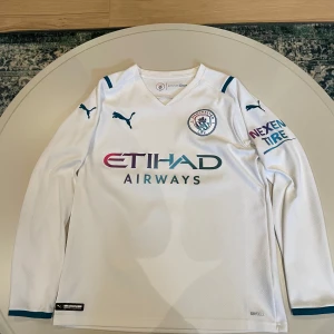 Manchester City 21/22 borta fotbollströja Puma  - Vit långärmad Manchester City fotbollströja från 21/22 Säsongen. Tröjan är ifrån Puma med klubbmärke på bröstet och sponsorloggor i blått och lila. Tillverkad i lätt och ventilerande Drycell-material, perfekt för träning eller match. Snygga blå detaljer på axlar och ärmslut. Tröjan är självklart äkta, den är köpt ifrån Classic football shirts. Tröjan är nyskick.