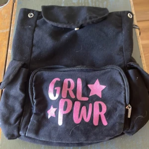 Svart ryggsäck med GRL PWR-tryck - Cool svart ryggsäck i bomull med stort fack och flera mindre fickor. Framsidan har ett rosa metallic GRL PWR-tryck med stjärnor. Praktisk dragkedja på ytterfacket och tryckknappar på sidorna. Perfekt för dig som vill sticka ut med girl power-vibe.
