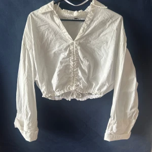 Vit croppad skjorta med snörning - Snygg vit croppad skjorta med lång ärm och krage. Skjortan har v-ringning och cool snörningsdetalj framtill samt resår i midjan för en chill passform.