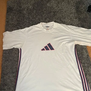 Vit Adidas t-shirt med ränder - Vit vintage t-shirt från Adidas med klassisk logga framtill i blått och rött med prickigt mönster. Snygga blå och röda ränder längs sidorna. Skön passform och rund halsringning.