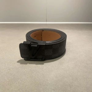 Louis Vuitton Damier svart bälte - Snyggt svart bälte från Louis Vuitton med klassiskt rutigt Damier-mönster och stor LV-logga i svart metall. Bältet är gjort i exklusivt skinn med brun insida och har en bred, modern look som sticker ut. Perfekt accessoar för att lyfta din outfit.