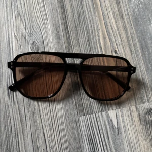 Svarta pilotbågar med bruna glas - Snygga solglasögon med svarta pilotbågar och bruna tonade glas. Klassisk och cool design med markerad ovandel och breda skalmar. Perfekta för att lyfta din stil med en retrovibe. Helt nya