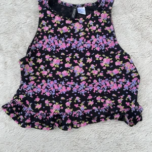 Blommig ärmlös topp från H&M Divided - Supersöt ärmlös topp från H&M Divided med svart botten och blommigt mönster i rosa, lila och lite grönt. Toppen har en croppad passform och volangkant nedtill som ger en extra gullig touch. Perfekt för dig som gillar färg och mönster!
