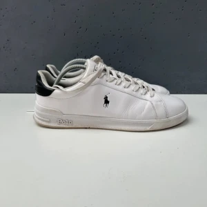 Vita sneakers från Polo Ralph Lauren - Snygga vita sneakers från Polo Ralph Lauren med svart detalj på hälen och klassisk logga på sidan. Skorna har snörning och är tillverkade i skinn med textilfoder och gummisula. Perfekta för en clean och stilren look.