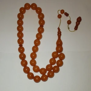 Orange pärlhalsband med stora kulor - Snygg Tasbih med stora, runda pärlor i en varm orange färg. Pärlorna är jämnt formade och sitter på en tunn guldfärgad tråd. Halsbandet har en unik detalj i form av en avlång pärla och dekorativa avslut. Perfekt för dig som vill sticka ut med färg.