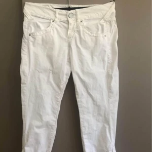 Vita byxor från Miss Sixty med knappar - Snygga vita byxor från Miss Sixty med coola detaljer som knappar längs bensluten och broderad gul fyrklöver baktill. Byxorna har låg midja, raka ben och är gjorda i bomull med lite stretch för skön passform. Perfekta för dig som gillar unik stil. 3/4 långa