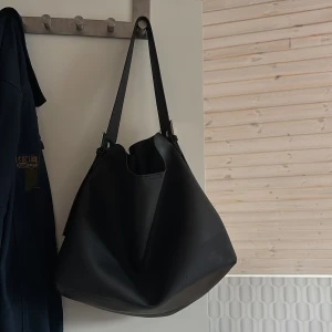 Svart axelväska i skinn med silverdetaljer - Stilren svart axelväska i slätt skinn med minimalistisk design. Väskan har bred axelrem med snygga silverfärgade spännen och rymligt huvudfack. Perfekt för dig som gillar enkel och modern look med plats för allt du behöver.