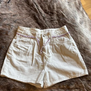 Vita högmidjade jeansshorts med snörning - Snörningen är virkad av mig, går att ta bort! Använd någon eller ingen gång av dottern! Ej stretch! Enligt mig för små för mig, jag är 169 cm och väger cirka 68-69kg! Snygga vita jeansshorts med hög midja och klassisk femficksdesign. Shortsen har en lila snörning i midjan som extra detalj och är tillverkade i ett stadigt bomullstyg. Perfekta för varma dagar och ger en fräsch look till din outfit.