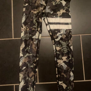 Stronger. Camo leggings med vita ränder - Snygga leggings från STRONGER i camomönster med vita ränder på ena benet. Tight passform och elastiskt material som passar perfekt till träning. Färgerna går i mörkgrönt, brunt, beige och svart. Midjan är normal och tyget är mjukt och stretchigt.