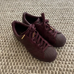 Vinröda Adidas Superstar  - Snygga Adidas Superstar i vinrött med fint äkta skinn och gulddetaljer på snörningen. Nästan aldrig använda och är lite dammiga. Skicket är mycket bra och de är väldigt bekväma. 