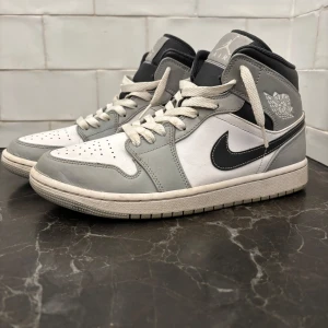 Nike Air Jordan 1 Mid grå/vit - Nike Air Jordan 1 Mid i stilren grå och vit färg med svarta detaljer. Klassisk high-top siluett med snörning och perforerad tå. Skorna har en svart Swoosh och ikonisk Air Jordan-logga på sidan. Tillverkade i skinn och textil för en clean och sportig look. Priset är ej hugget i sten.