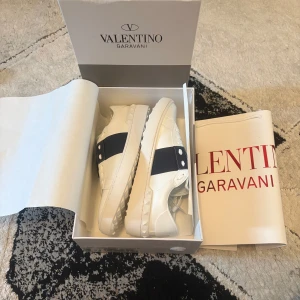 Valentino Garavani vita sneakers - Snygga vita sneakers från Valentino Garavani med bred svart rem över mitten och ikoniska nitar på hälen. Skorna har rund tå, platt sula och är tillverkade i skinn. Perfekta för dig som vill ha en clean men lyxig look.