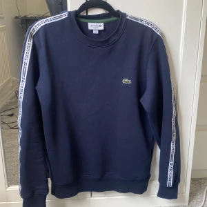 Mörkblå sweatshirt från Lacoste - Snygg mörkblå sweatshirt från Lacoste med klassisk krokodil-logga på bröstet och vita logoband längs ärmarna. Storleken är xs men passar på S. Köpte tröjan för 800kr har knappt användt den