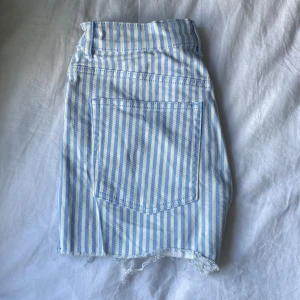 Blå randiga jeansshorts - Snygga blå- och vitrandiga jeansshorts med klassisk femficksmodell och knappstängning framtill. Perfekta för varma dagar och ger en fräsh sommarvibe. 