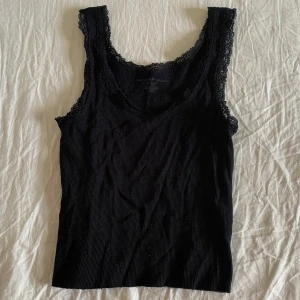 Svart spetskantad linnetopp Brandy Melville - Svart ribbad linnetopp från Brandy Melville med fina spetsdetaljer längs ringningen och axelbanden. Toppen är ärmlös och har en smickrande v-ringning. Tillverkad i mjuk bomull som känns skön mot huden. Perfekt att styla med jeans eller kjol.