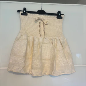 Beige kortkjol med smock från Zara - Supersöt beige kortkjol från Zara med smockad midja och knytband framtill. Kjolen har ett diskret vävt mönster och volangig nederdel som ger en luftig och chill vibe. Perfekt för varma dagar och lätt att matcha med crop tops eller linnen.