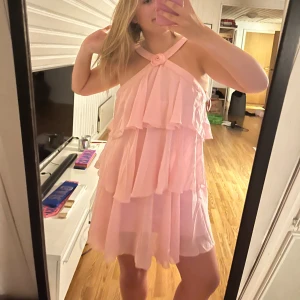 Rosa volangklänning med halterneck - Supersöt rosa kortklänning med volanger och halterneck med en liten rosdetalj framtill. Klänningen har flera lager och är ärmlös, perfekt för sommarens alla tillfällen. Lätt och luftig känsla. Endast använd en gång, köpt nu i Juni. Nypris 700kr