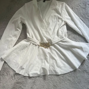 Vit omlottkavaj med kedjedetalj Amisu - Snygg vit blus från Amisu med lång ärm och markerad midja. Bältet har en cool guldfärgad kedjedetalj som ger extra edge. Skicket är som nytt.