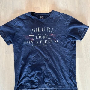 Blå t-shirt från Polo Ralph Lauren - Mörkblå t-shirt från Polo Ralph Lauren med tryck framtill i vitt och rött. Klassisk rund hals och korta ärmar. Tillverkad i 100% bomull, perfekt för en avslappnad och stilren look.