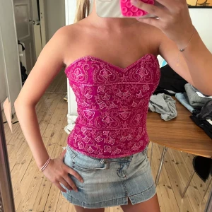 Rosa korsett med paisleymönster - Säljer en snygg rosa korsett med vitt paisleyinspirerat mönster. Toppen är axelbandslös och har en markerad sweetheart-ringning samt dragkedja i ryggen. Perfekt för dig som vill sticka ut med en färgstark och trendig look.