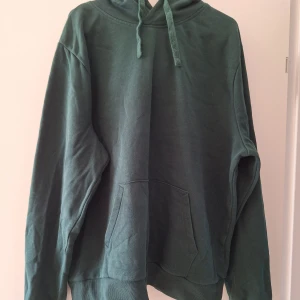 Mörkgrön hoodie från SOC i bomull - Mörkgrön hoodie från SOC i storlek M. Klassisk modell med huva och snörning, samt stor magficka framtill. Tillverkad i mjuk bomulls- och polyestermix som är skön att ha på sig. Perfekt för chill dagar eller när du vill ha en enkel och stilren look.