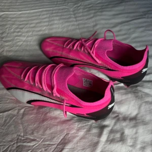 Puma Ultra rosa fotbollsskor - Snygga Puma Ultra fotbollsskor i en riktigt poppig rosa färg med svarta och vita detaljer. Skorna har snörning, låg profil och en lätt ovandel i syntetmaterial. Sulan har dobbar för gräsplan och coola loggor på hälen och undersidan. 