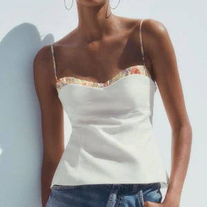 Vit blommig linnetopp med smala axelband - Supersöt vit linnetopp med smala axelband och blommigt mönster längs kanten och banden. Sitter ganska tajt så skulle nog säga att den sitter mer som en XS, helt oanvänd och slutsåld på hemsidan!