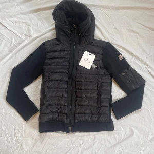 Moncler cardigan svart med huva  - Snygg svart pufferjacka från Moncler med stickade ärmar och quiltad front. Jackan har huva, dragkedja framtill och en extra ficka med dragkedja på ärmen. Perfekt för dig som vill ha en stilren och exklusiv look. Den är helt ny och aldrig använd och är i mycket bra skick och kvalité.