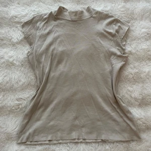 Beige polotopp i bomull - Säljer en enkel och stilren beige polotopp med korta ärmar. Toppen har en högre krage och är gjord i mjuk bomull, perfekt för en clean och avslappnad look. Passformen är normal och toppen är lätt att matcha med olika outfits.