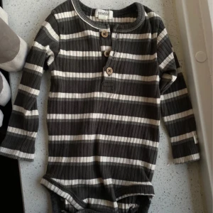 Randig ribbad body från Newbie, barn, 68 - Superfin långärmad body från Newbie i ribbad bomull. Mörkgrå och vita ränder, tre snygga träknappar framtill och mjuk känsla. Perfekt för dig som gillar stilrena och bekväma plagg till de små.