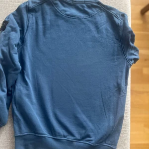 Blå Stone Island sweatshirt - Säljer en blå sweatshirt från Stone Island med klassisk rund hals och lång ärm. Tröjan har den ikoniska Stone Island-loggan på vänster ärm. Tillverkad i mjuk bomull och har en clean, stilren look som passar perfekt till jeans eller joggers.