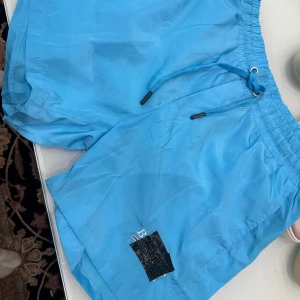 Blåa shorts från Balenciaga - Snygga blåa shorts från Balenciaga med elastisk midja och snörning. Shortsen har en klassisk passform och är tillverkade i ett lätt syntetmaterial, perfekta för sommaren. Detalj med broderad logga på benet, delvis täckt. Stilrena och sportiga vibes.