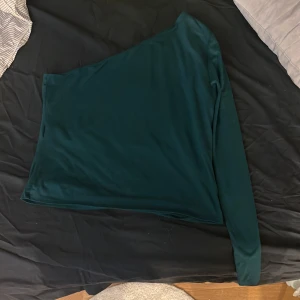 Mörkgrön one shoulder topp H&M - Säljer en mörkgrön one shoulder topp från H&M i storlek L. Toppen har lång ärm på ena sidan och ärmlös på andra, vilket ger en snygg asymmetrisk look. Perfekt för dig som vill sticka ut lite extra med en stilren och trendig topp. !köpare står för frakt!