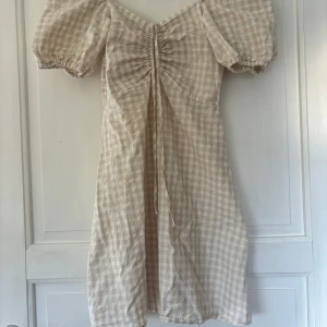 Beige rutig kortklänning H&M XS - Supersöt kortklänning från H&M i beige och vitt rutigt mönster. Klänningen har puffärmar, smock i ryggen och snörning framtill. Endast använd en gång💞